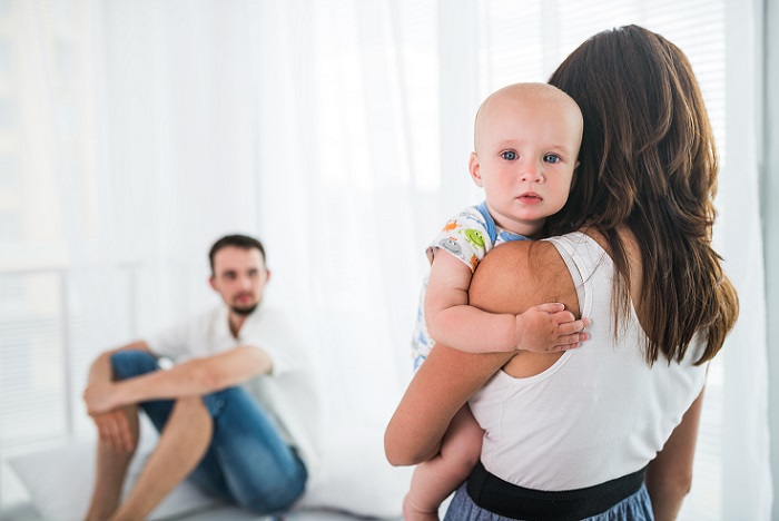 Unidentified young woman holds a small child in her arms on the background of a sitting upset, blurred young man. Family breakdown and divorce concept